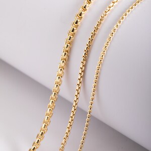 May include: Three gold-toned box chains of varying thicknesses arranged side by side. Each chain features a series of interconnected square links with a polished, reflective finish, showcasing a consistent design across all three strands.