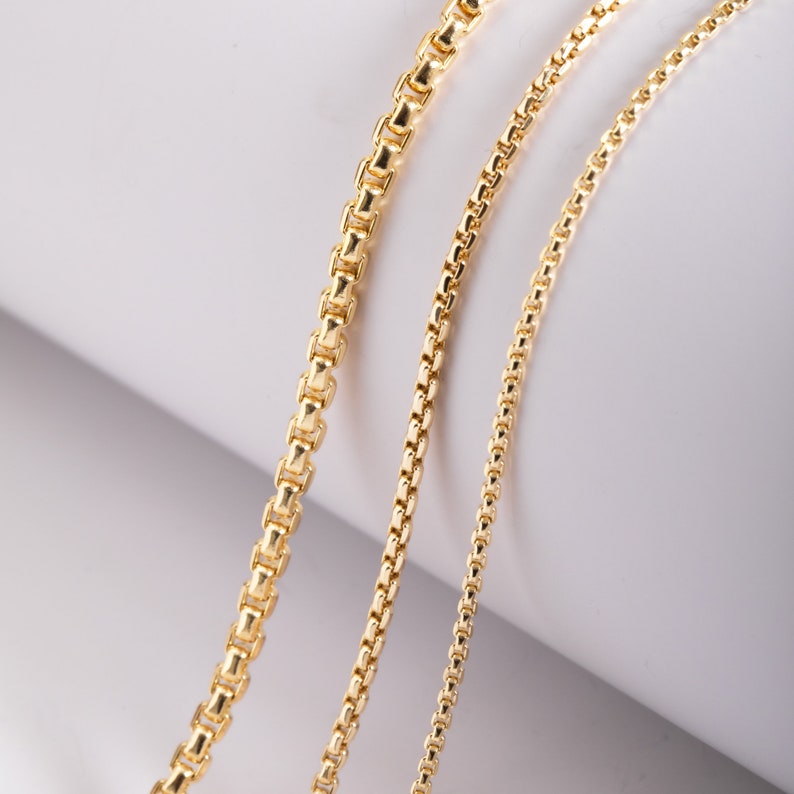 May include: Three gold-toned box chains of varying thicknesses arranged side by side. Each chain features a series of interconnected square links with a polished, reflective finish, showcasing a consistent design across all three strands.