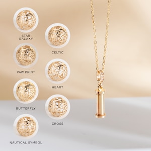May include: A gold necklace with a cylindrical pendant and a variety of gold charms with different designs, including a star galaxy, celtic, paw print, heart, butterfly, cross, and nautical symbol.
