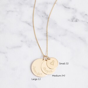 May include: A gold-colored necklace with three circular pendants of varying sizes. The largest pendant is engraved with the name "Jennifer", the middle one with "Shopie", and the smallest with a heart symbol. The necklace is displayed against a marble background.