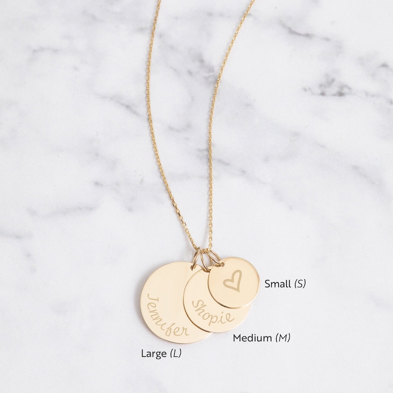 May include: A gold-colored necklace with three circular pendants of varying sizes. The largest pendant is engraved with the name "Jennifer", the middle one with "Shopie", and the smallest with a heart symbol. The necklace is displayed against a marble background.
