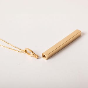 Op de afbeelding: Een goudkleurige, rechthoekige hangerketting met een schroefdop. De hanger is losgemaakt van de ketting, waardoor de schroefdraadopening zichtbaar is. De ketting en hanger zijn gemaakt van glanzend, goudkleurig metaal. De ketting wordt op een wit oppervlak getoond.