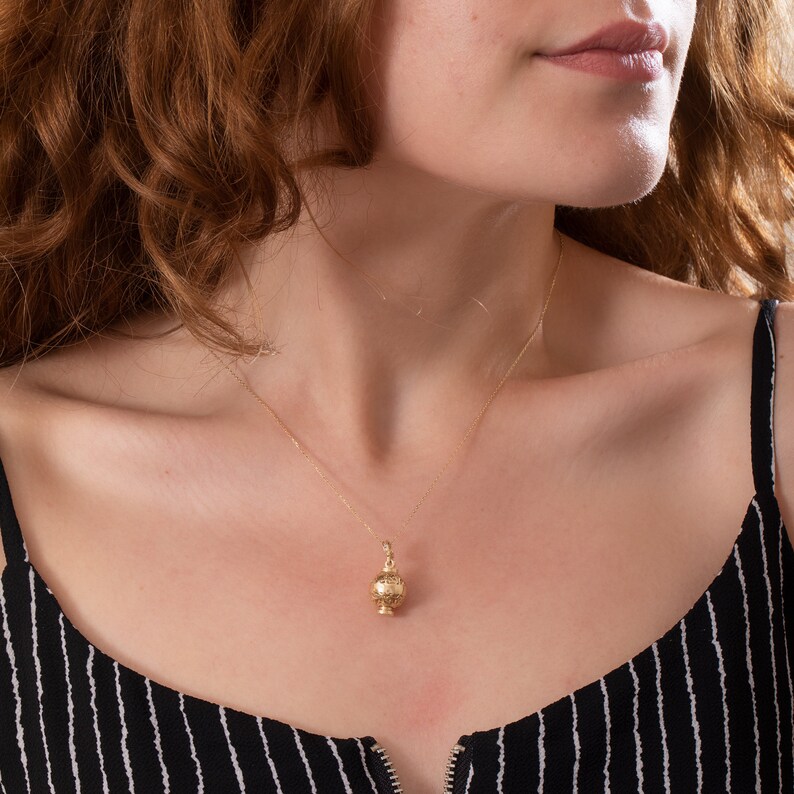 May include: A gold necklace with a delicate, ornate pendant in the shape of a small, round vase. The necklace is worn by a person with long, curly red hair.