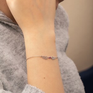 Puede incluir: Una delicada pulsera de plata con dijes en forma de alas a cada lado de una peque&ntilde;a gema roja redonda. La pulsera se lleva en una mu&ntilde;eca, con un su&eacute;ter gris en el fondo.
