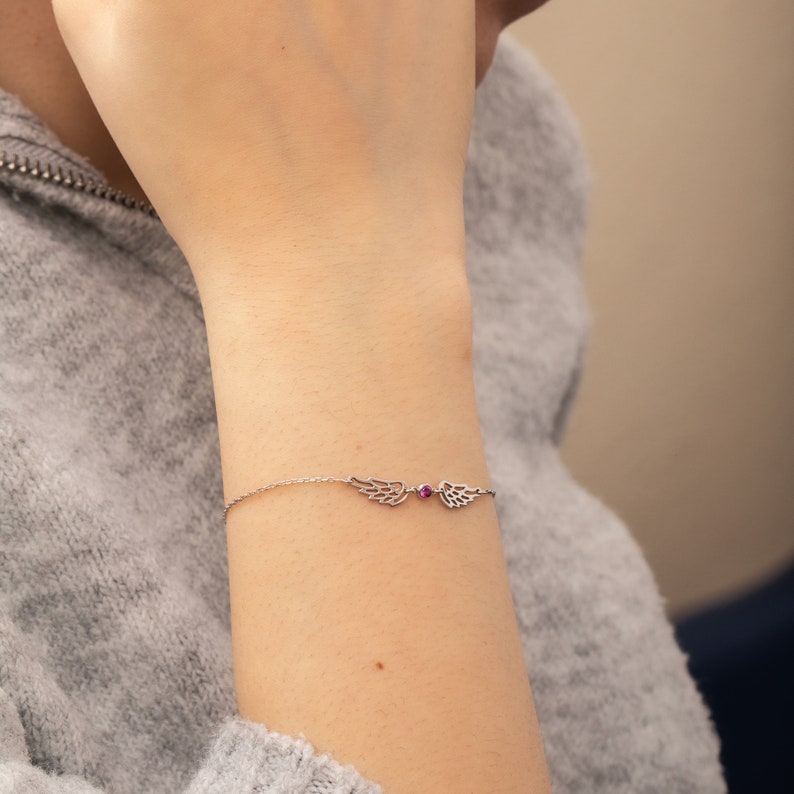 Puede incluir: Una delicada pulsera de plata con dijes en forma de alas a cada lado de una peque&ntilde;a gema roja redonda. La pulsera se lleva en una mu&ntilde;eca, con un su&eacute;ter gris en el fondo.