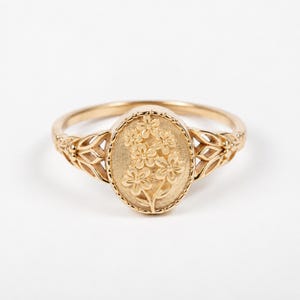 May include: A gold ring featuring an oval-shaped centrepiece with an embossed floral design. The band displays intricate cut-out leaf-like patterns on both sides, leading to a smooth, polished finish that encircles the finger with a delicate aesthetic.