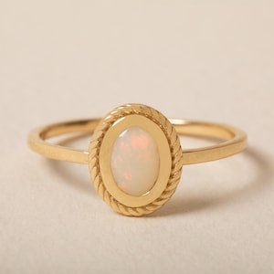 May include: A gold ring featuring an oval-shaped opal gemstone set in a bezel. The setting is surrounded by a decorative twisted rope-style border. The band is thin, smooth, and polished, creating a refined look for this piece of jewelry.