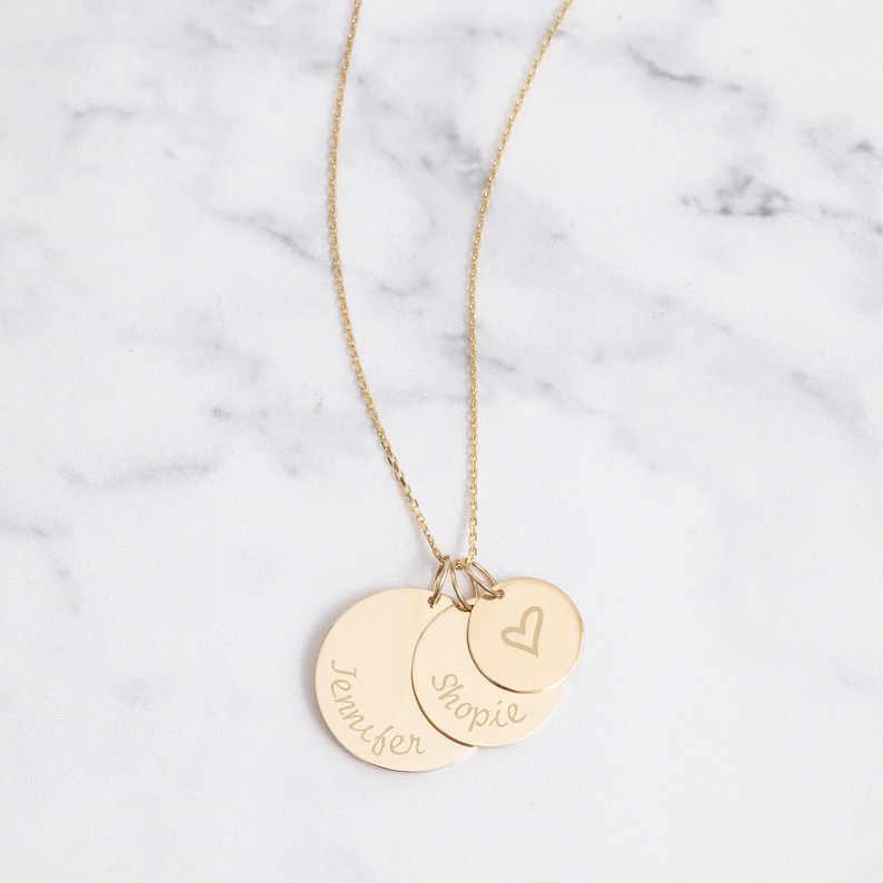 May include: A gold-colored necklace with three circular pendants. The largest pendant has the name "Jennifer" engraved on it. The middle pendant has the name "Shopie" engraved on it. The smallest pendant has a heart symbol.