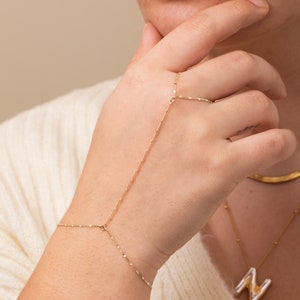 May include: A gold chain bracelet with a delicate design. The bracelet is connected to a ring that can be worn on the finger. The bracelet is shown on a person's hand.