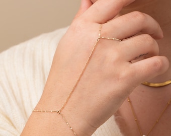 May include: A gold chain bracelet with a delicate design. The bracelet is connected to a ring that can be worn on the finger. The bracelet is shown on a person's hand.