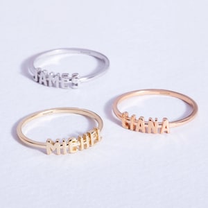 Puede incluir: Tres anillos de oro con nombres escritos en letras de bloque. Los anillos están sobre una superficie blanca. Los nombres son "James", "Liana" y "Michael".