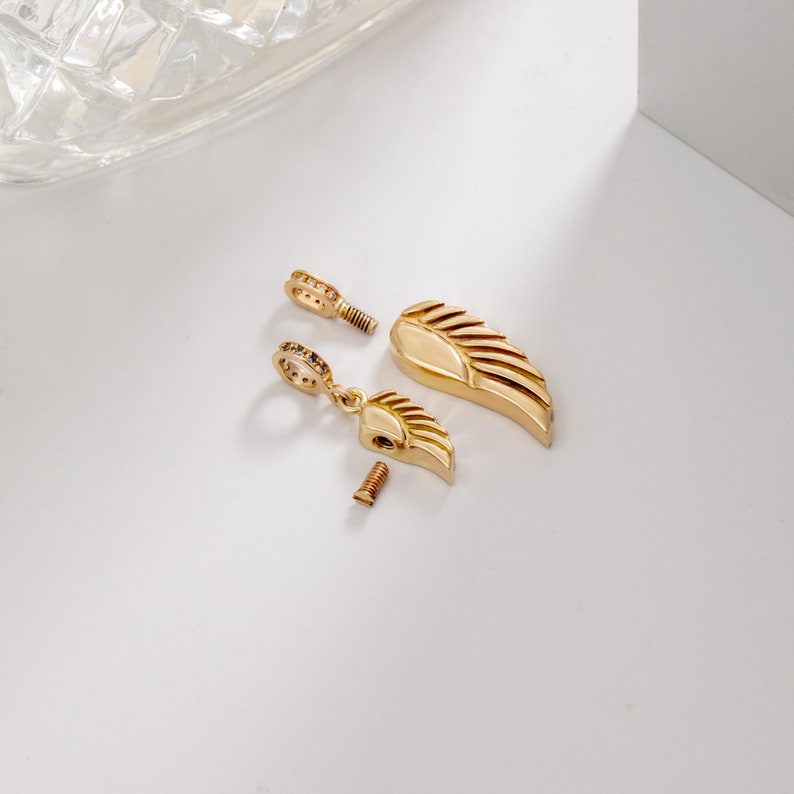 K&ouml;nnte beinhalten: Goldfarbener Engel Fl&uuml;gel Krematoriumsanh&auml;nger mit einem Schraubverschluss und einem kleinen, mit Kristallen besetzten Ring. Der Anh&auml;nger ist zerlegt und zeigt die Schraube und den Ring.