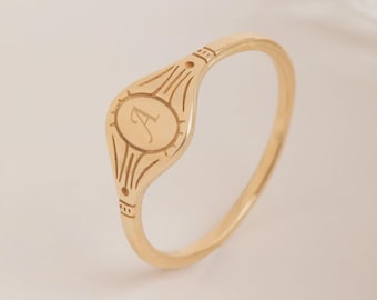 May include: A gold-toned signet ring featuring a smooth, oval-shaped face engraved with the letter A in a cursive script. The face is surrounded by etched sunburst-style lines that extend onto the thin, polished band of the ring.