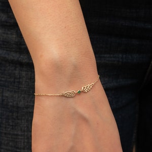 Puede incluir: Una delicada pulsera dorada con dos colgantes de alas estilizadas. Una peque&ntilde;a gema verde est&aacute; centrada entre las alas. La pulsera se muestra en una mu&ntilde;eca sobre un fondo oscuro.