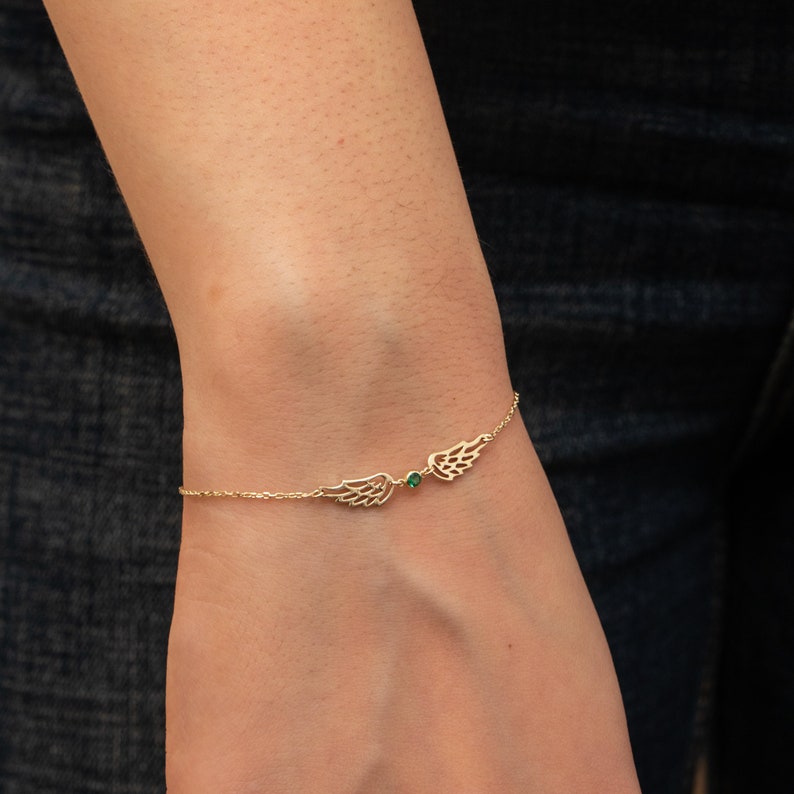 Puede incluir: Una delicada pulsera dorada con dos colgantes de alas estilizadas. Una peque&ntilde;a gema verde est&aacute; centrada entre las alas. La pulsera se muestra en una mu&ntilde;eca sobre un fondo oscuro.