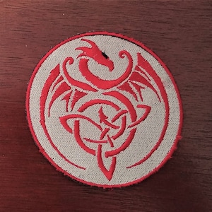May include: A red embroidered dragon design on a white circular patch. The dragon is curled up in a knot with its wings spread out.
