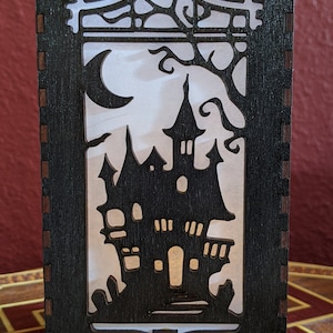 May include: A black wooden lantern with a cutout design of a spooky house and a crescent moon. The lantern is lit from within, casting a warm glow.