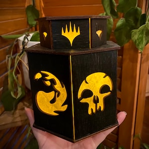 May include: A black wooden hexagonal box with a light-up design. The box features a skull, a dragon, and the Magic: The Gathering symbol.