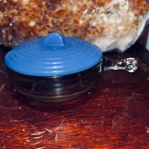 May include: A small, clear glass dish with a blue lid. The dish has a handle and is sitting on a wooden surface. A large, rough, orange and brown crystal formation is in the background.