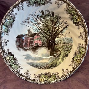 May include: A decorative ceramic bowl featuring a rural landscape scene. The bowl's interior depicts a red barn, a large tree, and a winding road. The rim is decorated with a black and white floral pattern with green accents.