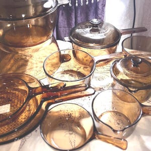 Puede incluir: Una colección de utensilios de cocina de vidrio color ámbar que incluye una olla grande con tapa, varias cacerolas con tapas y sartenes abiertas. Cada pieza presenta un tono marrón translúcido y una construcción de vidrio liso con asas integradas.