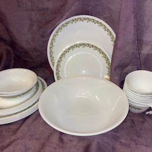 以下が含まれることがあります： 緑色の花の縁取りが付いた白い食器セット。セットには、お皿、ボウル、カップが含まれています。お皿とボウルは積み重ねられ、カップはきちんと並べられています。背景は紫です。