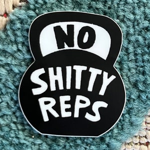 May include: Black and white sticker in the shape of a kettlebell. The sticker has the words "NO SHITTY REPS" in bold white letters. The sticker is on a textured blue and white surface.