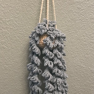 May include: A handmade, gray crochet plant hanger with a cream-colored hanging cord. The plant holder is a cascading design, with a beige ball at the top. The crochet work has a textured, floral-like pattern.
