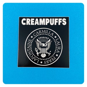 May include: Black and white sticker with the text "CREAMPUFFS" and a seal with the text "CARMILLA LAURA PERRY LAFONTAINE" around a stylized eagle.