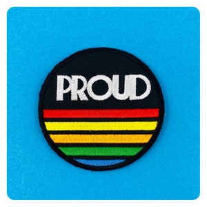 May include: A black and white embroidered patch with the word "PROUD" in white letters on a black background. The patch has a rainbow stripe design with red, orange, yellow, green, blue, and purple stripes.