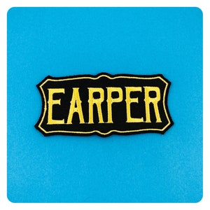 Pode incluir: Etiqueta bordada preta e amarela com a palavra "EARPER" em uma fonte decorativa.