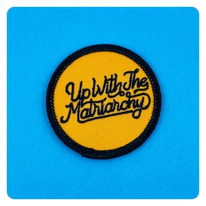May include: A yellow circle patch with black stitching around the edge. The text "Up With The Matriarchy" is embroidered in black on the patch.