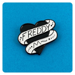 Può includere: Spilla in smalto nero e bianco con un cuore con uno stendardo che dice "FREDDY".