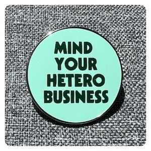 May include: A round, light blue enamel pin with a silver border. The pin says "MIND YOUR HETERO BUSINESS" in dark green letters.
