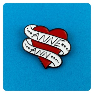 Può includere: Un cuore rosso con uno striscione bianco avvolto attorno. Lo striscione dice "ANNE" in alto e "ANN" in basso.