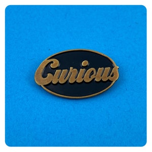Op de afbeelding: Een ovale gouden pin met een zwarte achtergrond. Het woord "Curious" is geschreven in een gouden schrijfletter.
