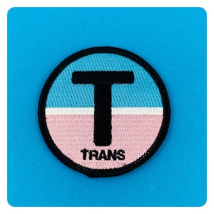 Puede incluir: Un parche bordado en blanco y negro sobre un fondo azul y rosa. El parche presenta una gran letra negra "T" con la palabra "TRANS" debajo.