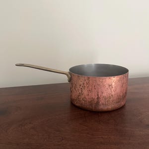 May include: A round copper saucepan with a weathered, textured exterior finish and a long, straight brass handle. The interior of the pan is smooth and metallic silver in colour, featuring a flat base and straight vertical sides.