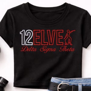 May include: A black crop top with the text "12 ELVE Delta Sigma Theta" is paired with light blue denim shorts, a black belt, and red, black, and white sneakers. Accessories include a black purse, sunglasses, and a black water bottle.