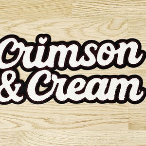 May include: A white and maroon patch with the words "Crimson & Cream" in a cursive font. The patch has a black border and a small heart above the letter "o" in "Crimson". The patch is on a light wood surface.