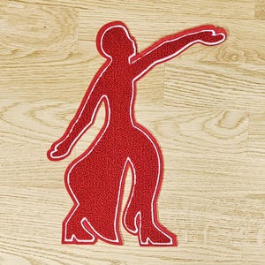 May include: A red rug in the shape of a person in a dancing pose, with white trim outlining the figure. The rug is placed on a light brown wooden floor. The figure has one arm extended and the other bent.
