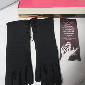 Pode incluir: Um par de luvas pretas Isotoner com tr&ecirc;s pequenos bot&otilde;es no punho. As luvas s&atilde;o exibidas ao lado de um cart&atilde;o promocional que diz "the ISOTONER Glove" e uma caixa com o texto "ARIS GLOVES, INC."