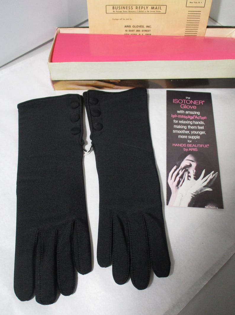 Pode incluir: Um par de luvas pretas Isotoner com tr&ecirc;s pequenos bot&otilde;es no punho. As luvas s&atilde;o exibidas ao lado de um cart&atilde;o promocional que diz "the ISOTONER Glove" e uma caixa com o texto "ARIS GLOVES, INC."