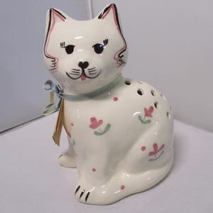 May include: A white ceramic cat figurine with black and pink accents. The cat has a blue ribbon around its neck and is decorated with pink flowers and dots. The figurine has holes on its back.