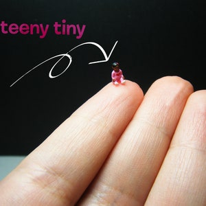 May include: A miniature figurine of a baby in a pink outfit, sitting on a fingertip. The figurine has dark hair and is wearing a pink onesie. The words "teeny tiny" are written above the figurine.