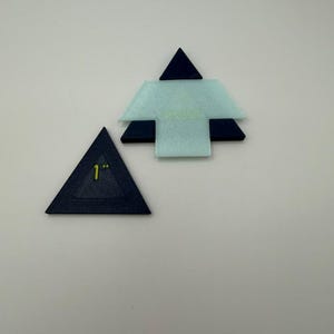May include: Two flat, geometric crafting templates. One is a dark blue triangle with the text 1 inch printed in yellow. The other is a layered assembly featuring a light blue, translucent, T-shaped piece centered over a dark blue triangle base.