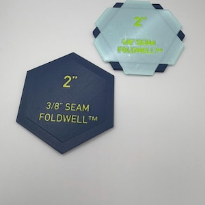 Puede incluir: Dos plantillas de costura hexagonales, una azul oscuro y otra azul claro translúcido, cada una con el texto 2" y 3/8" SEAM FOLDWELL™. Las plantillas están diseñadas para doblar tela y marcar márgenes de costura con medidas impresas.