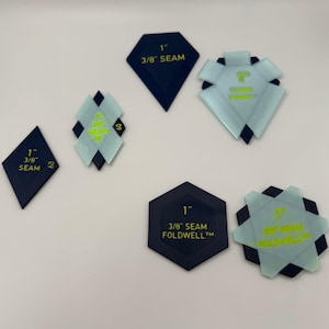 May include: A collection of dark blue and translucent light blue geometric quilting templates. Each piece features printed text including 1 inch, 3/8 inch seam, and Foldwell branding. The shapes include diamonds, hexagons, and pentagons.