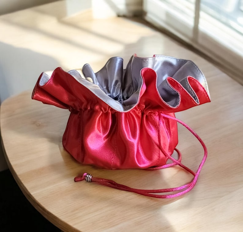 Op de afbeelding: Een roze satijnen trekkoordtasje met een zilveren voering. Het tasje is open en het trekkoord is zichtbaar.