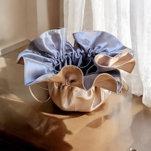 Op de afbeelding: Een licht gouden satijnen trekkoordtas met een lichtblauwe satijnen voering. De tas is open en de voering is zichtbaar. De tas ligt op een houten oppervlak.